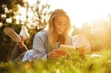 Bir kitap okuma memnun ile genç bir kız parkta bir çim üzerinde döşeme sırt çantası portresi
