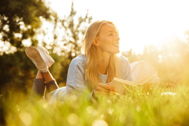 Bir kitap okuma memnun ile genç bir kız parkta bir çim üzerinde döşeme sırt çantası portresi