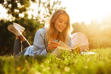 Bir kitap okuma gülümseyen genç bir kız parkta bir çim üzerinde döşeme sırt çantası ile portresi