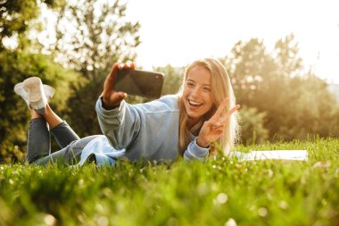 Selfie, barış gösterilen alarak güzel bir genç kız parkta, çimlerin üzerine döşeme kitap ile portresi