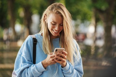 Cep telefonu tutan bir gülümseyen genç kız öğrenci parkta, yürüyüş sırt çantası ile kulaklık portresi