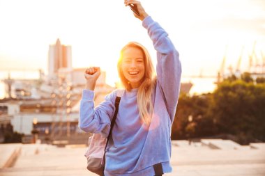 Sırt çantası ayakta açık havada sırasında günbatımı, holding cep telefonu, Genç ve güzel bir kızla portresi kutluyor