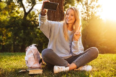 Selfie, barış gösterilen alarak güzel bir genç kız bir ot parkta oturan sırt çantası ile portresi