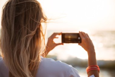 Cep telefonu ile sahilde bir gün batımı bir resim çekmek bir kadının arkadan görünüm