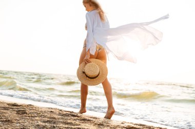 Plaj boyunca yürüme çekici genç bir kadın şapkası, elinde yaz giysileri resim kırpılmış