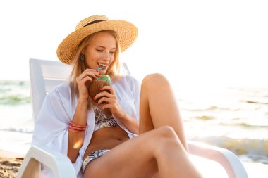 Görüntü muhteşem beyaz kadının 20s sırasında yaz sabah sahilde şezlong içinde otururken tatlı kokteyl içme hasır şapkalı