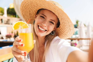 Güzel genç kız yaz şapka ve güneşli kumsalda dinlenme, bir kokteyl ile bir kafede oturan, bir selfie alarak mayo