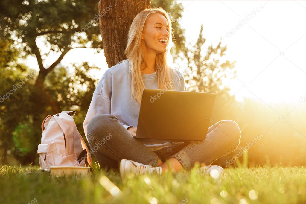 Portrait of a cheerful young girl with backpack sitting with legs crossed, using laptop computer