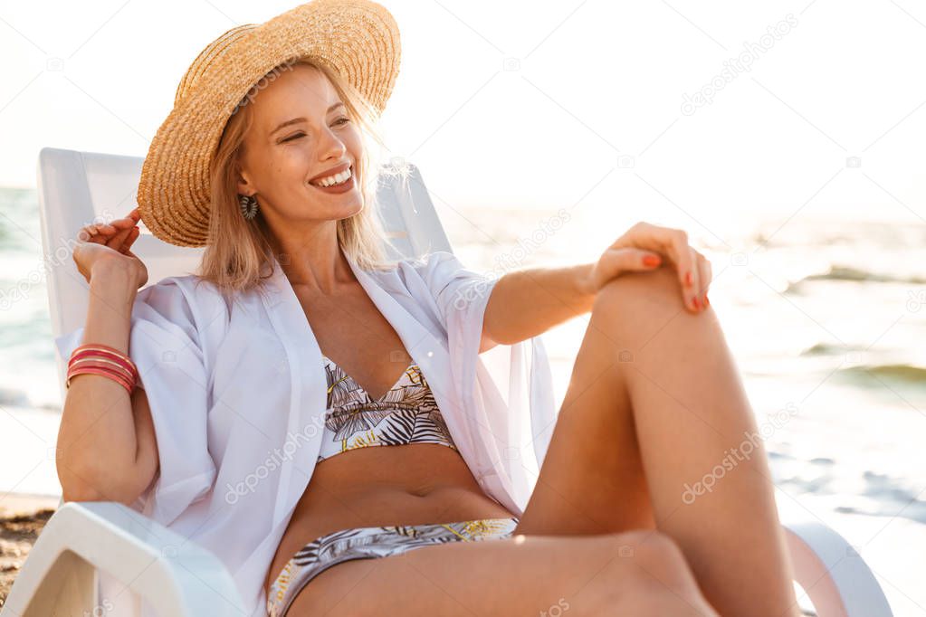 Photo of pretty blonde woman 20s in swimsuit and straw hat smiling while lying in deck chair at sea coast during summer sunrise