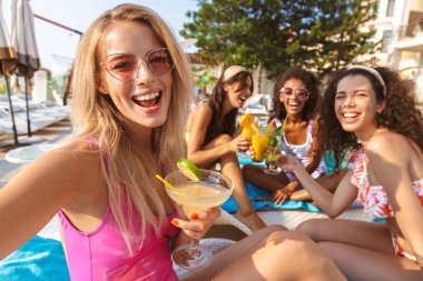 Mutlu duygusal grup havuzda açık havada oturup kokteyl içme kadın arkadaşıyla swimwears içinde giyinmiş genç görüntüsünü almak bir selfie kamera tarafından.