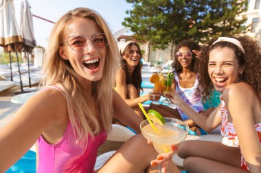 Mutlu duygusal grup havuzda açık havada oturup kokteyl içme kadın arkadaşıyla swimwears içinde giyinmiş genç görüntüsünü almak bir selfie kamera tarafından.