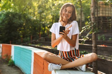 Genç kız müzik kulaklık ve vasıl belgili tanımlık park açık havada hareket eden telefon ile gülümseyen