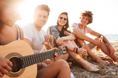 Grup neşeli genç arkadaşlar eğlenmeye zaman birlikte sahilde bira içme, kamp sırasında gitar çalmak,