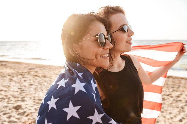 Image of cheerful excited women friends outdoors on the beach holding USA flag having fun.