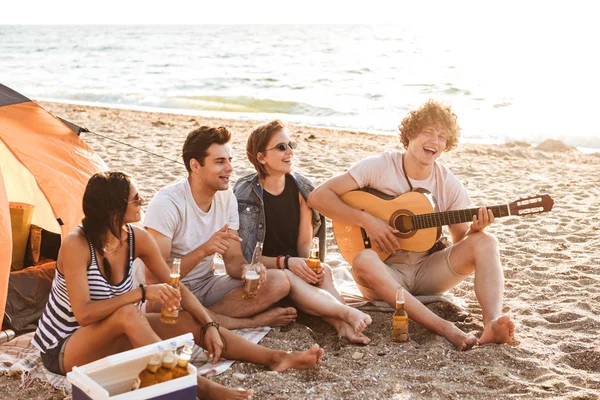Group of excited young friends having fun time together at the beach, drinking beer, playing guitar while camping