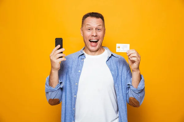 一个兴奋的中年男子的肖像使用手机 持有信用卡隔离在黄色背景 — 图库照片
