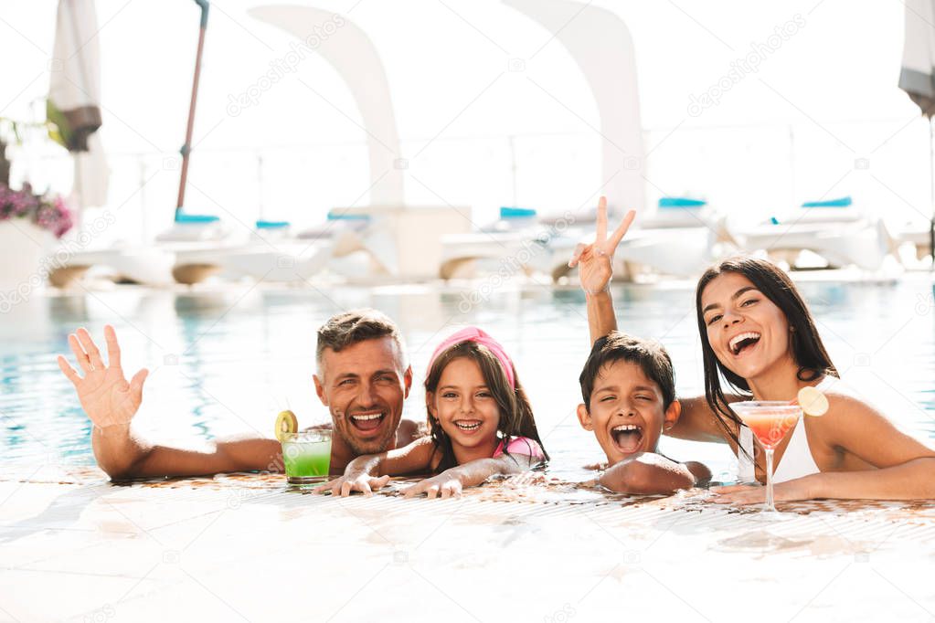 Happy young family having fun inside a swimming pool outdoors in summer with cocktails, waving hands