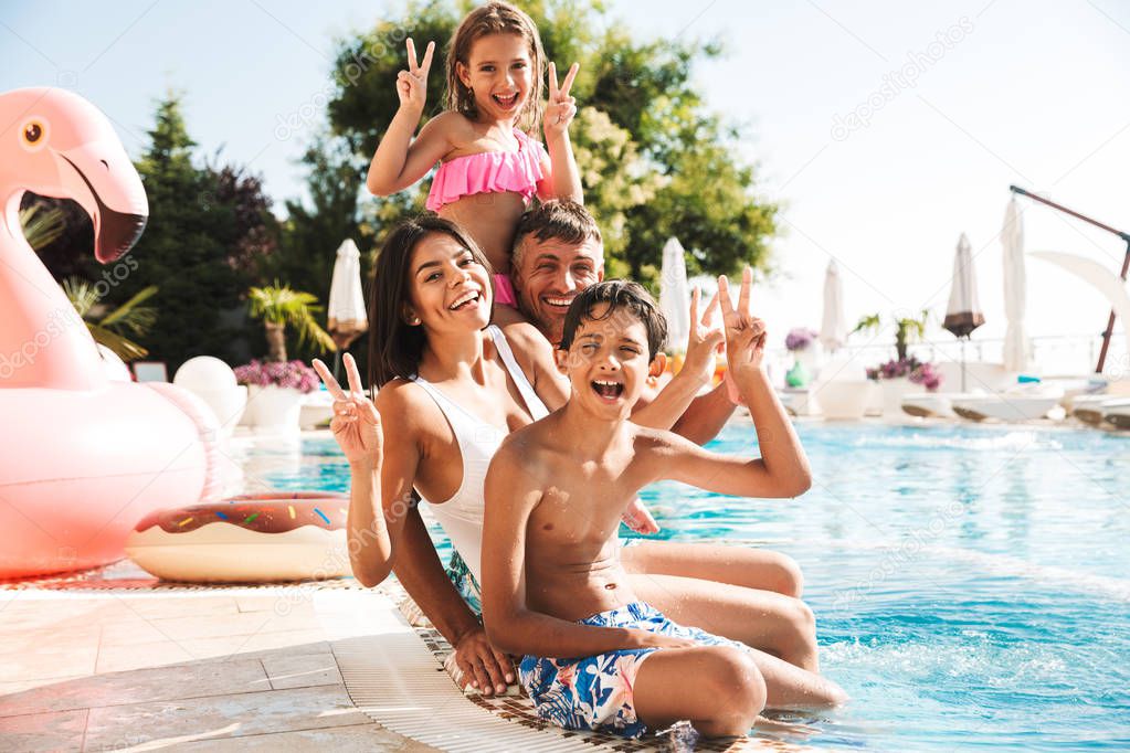 Happy young family having fun together at the swimming pool outdoors in summer, showing peace gesture