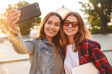 Mutlu genç güzel bayanlar öğrenciler parkta yürüyüş görüntüsünü almak bir selfie cep telefonuyla.