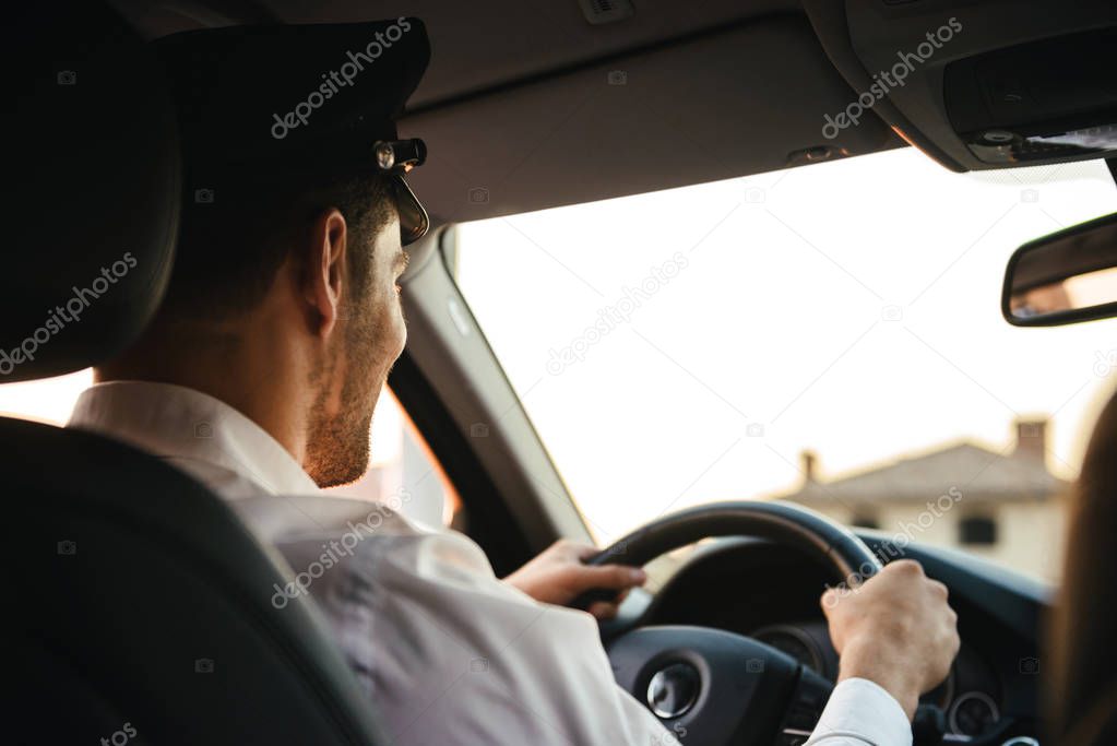 Portrait from back of caucasian chauffeur man wearing uniform and cap holding wheel and driving car