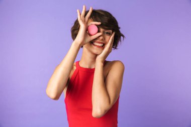 Görüntü gülümseyen ve menekşe arka plan üzerinde izole macaron bisküvi tutarak rahat kıyafet içinde mutlu güzel kadın 20'li