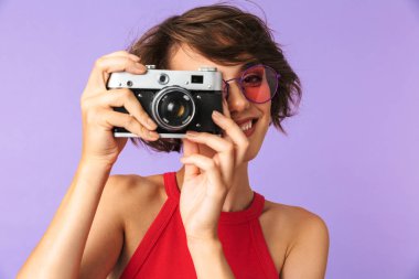 Hippi kız 20'li yaşlarda gülümseyen ve menekşe arka plan üzerinde izole retro kamerada fotoğraf çekme güneş gözlüğü fotoğrafı