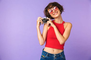 Mutlu kadın 20'li yaşlarda gülümseyen ve menekşe arka plan üzerinde izole retro kamerada fotoğraf çekme güneş gözlüğü