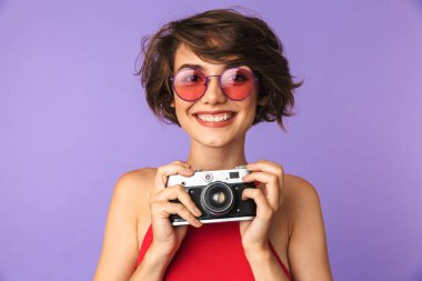 Trendy kız 20'li yaşlarda gülümseyen ve menekşe arka plan üzerinde izole retro kamerada fotoğraf çekme güneş gözlüğü fotoğrafı