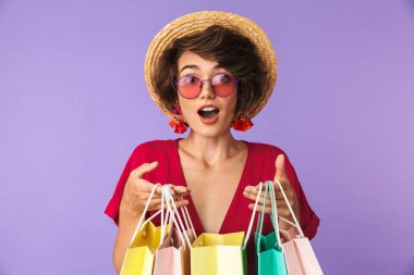 Image of excited brunette woman 20s in straw hat holding colorful paper shopping bags isolated over violet background