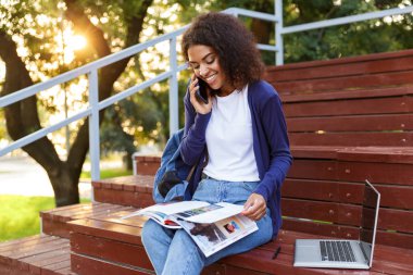 Dergi okuma bir neşeli genç Afrikalı kız parkta, istirahat sırasında cep telefonu üzerinde konuşurken sırt çantası ile portresi