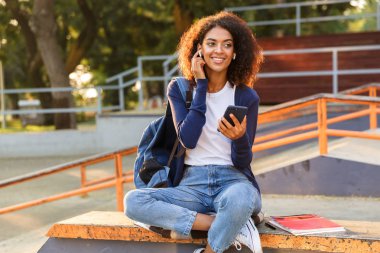 Açık havada cep telefonu dinleme müzik kulaklık kullanarak parkta oturan mutlu Afrika genç kadın görüntüsünü.