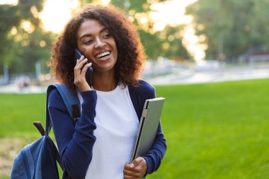 Görüntü genç güzel Afrikalı kadın öğrencinin cep telefonuyla konuşurken laptop tutan parkta yürüyüş.