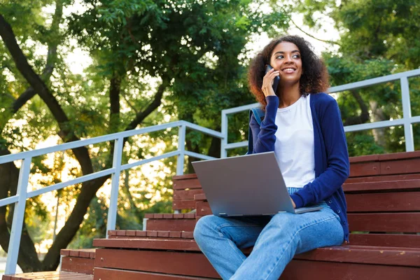 Açık havada cep telefonuyla konuşurken dizüstü bilgisayar kullanan basamaklarda parkta oturan mutlu Afrika genç kadın görüntüsünü.