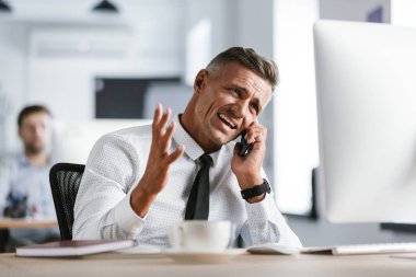 Tahriş olmuş işadamı 30'lu giyen beyaz gömlek ve kravat Office bilgisayar ve smartphone üzerinde konuşurken çığlık masada oturan görüntü