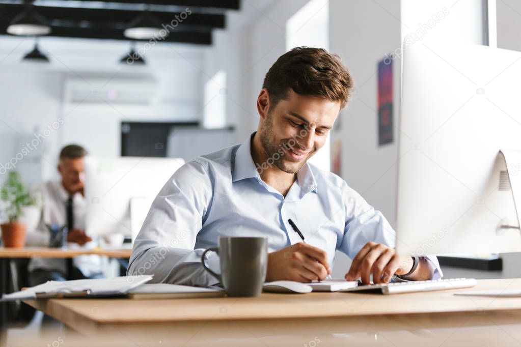 Image of Smiling male manager writing something while sitting by the table in office