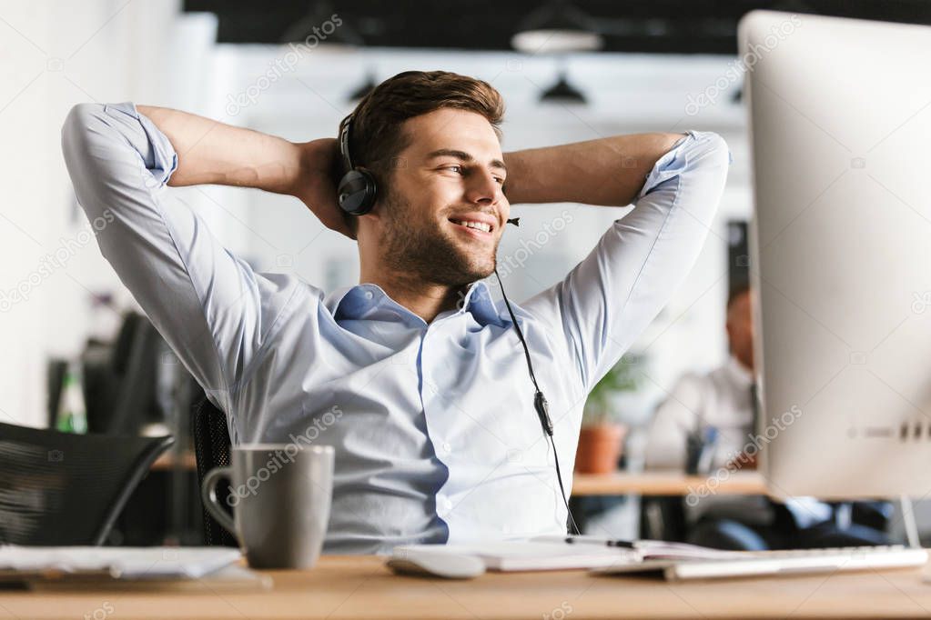 Pleased office manager talking by headset with microphone and looking away while sitting by the table in office