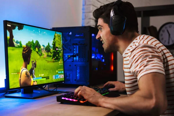 Portrait of caucasian gamer guy looking at screen while playing video games on computer wearing headphones and using backlit colorful keyboard
