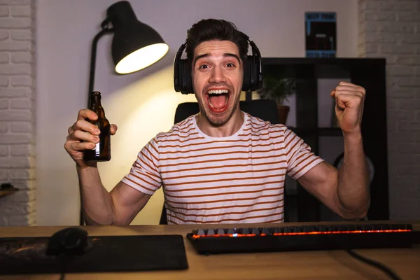 Portrait of joyful gamer guy wearing headphones screaming and drinking beer while sitting at desk with computer in room