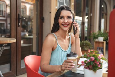 Kafede açık havada kahve içme cep telefonuyla konuşurken oturan genç neşeli kadın görüntüsünü.