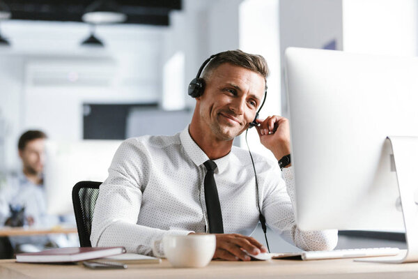 Photo of joyful operator man 30s wearing office clothes and headset smiling while working on computer in call center