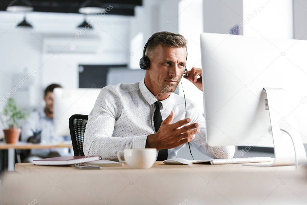 Photo of businesslike man 30s wearing office clothes and headset sitting by computer in call center