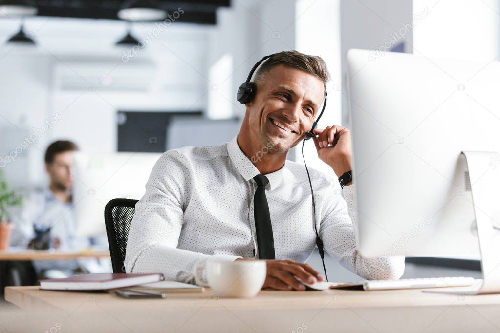 Photo of smiling operator man 30s wearing office clothes and headset sitting by computer in call center