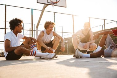 Neşeli çok ırklı gençler basketbol oyuncu oturma grubu spor yere konuşuyor, dinlenme