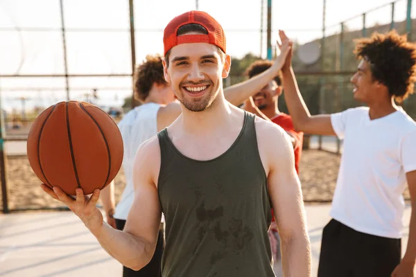 Portrait of caucasian muscular guy spinning ball on his finger while playing basketball at the playground outdoor with his friends