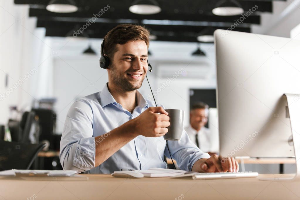 Photo of businessman 30s wearing office clothes and headset drinking tea from cup while sitting by computer in call center