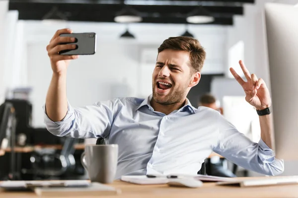 Playful business man making selfie on smartphone and showing peace gesture while sitting by the table in office