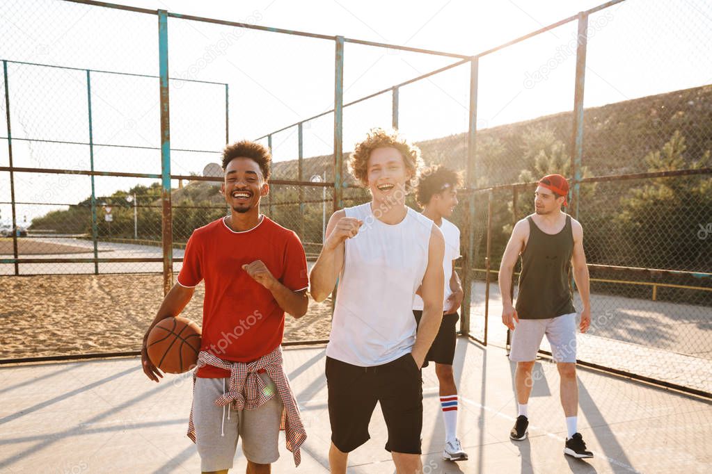 Photo of young sporty men smiling and holding ball while standing at basketball playground outdoor during summer sunny day