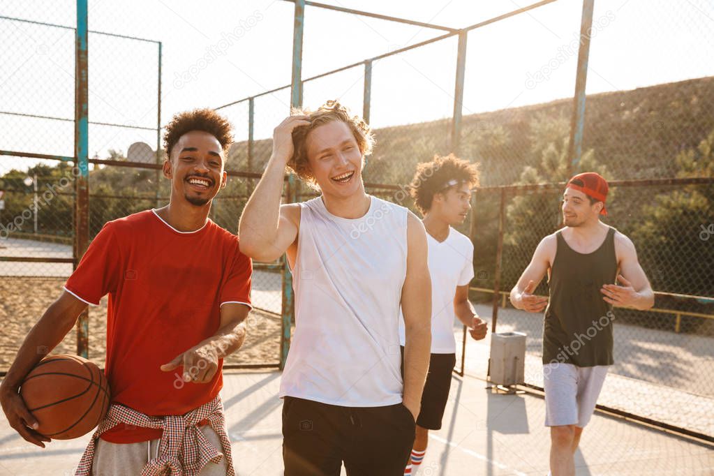 Photo of muscular sporty men smiling and holding ball while standing at basketball playground outdoor during summer sunny day