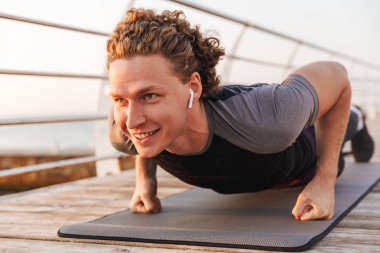 Smiling young sportsman in wireless earphones doing push ups on a fitness mat outdoors clipart