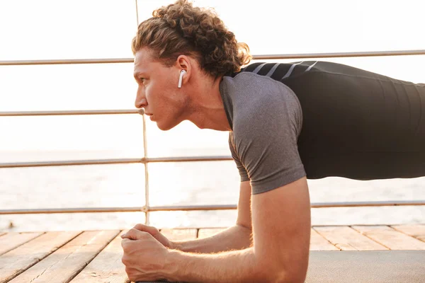 一个强壮的运动员的肖像做木板运动在健身垫海滩 听音乐与耳机 — 图库照片